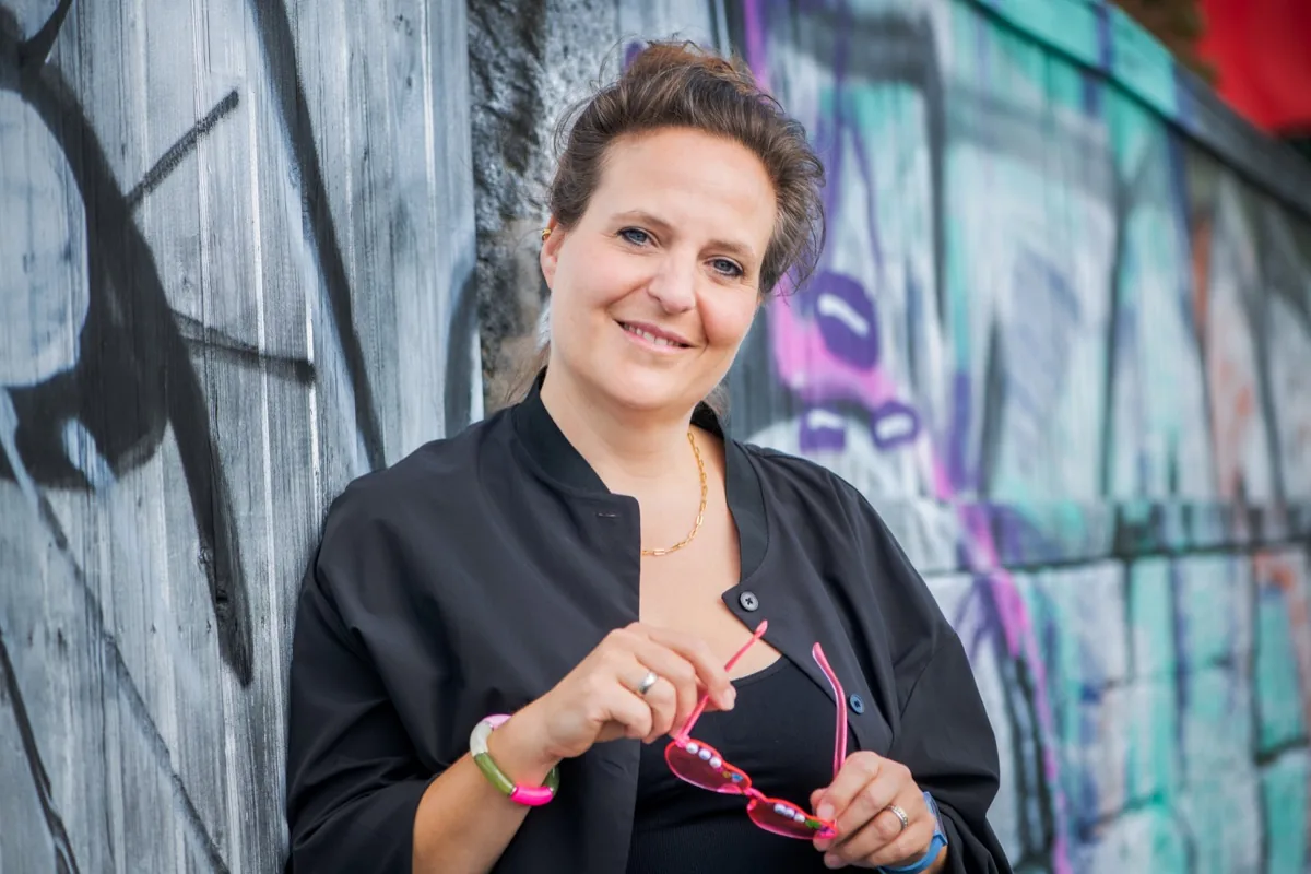 Freie Rednerin Annette Krönert lächelt mit rosa Brille in der Hand vor einer bemalten Wand